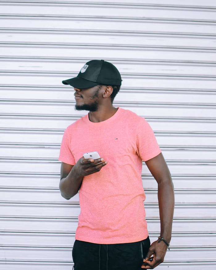 Man wearing a pink shirt holding a smartphone, looking aside against a striped backdrop.