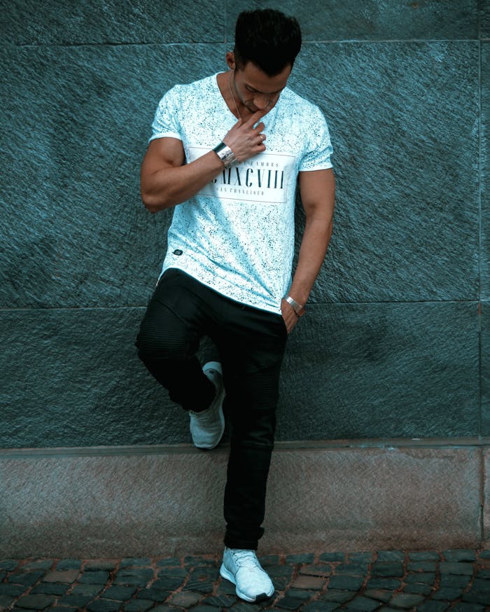 Stylish young man in casual attire leaning against a city wall, exuding a pensive mood.
